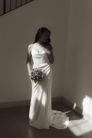 A bride in an elegant white dress poses with her bouquet in dramatic window light.
