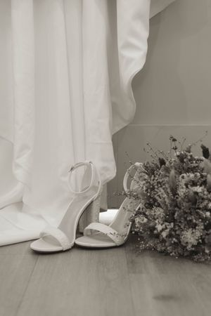 Close-up of white bridal shoes and dried flower bouquet against flowing fabric.