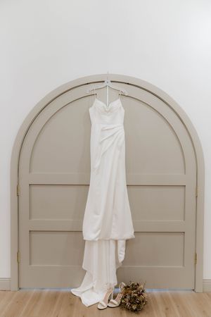 Elegant white wedding dress hanging on arched doorway with dried flower bouquet below.