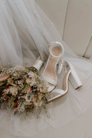 Wedding accessories including white heels and delicate floral arrangements on light fabric.