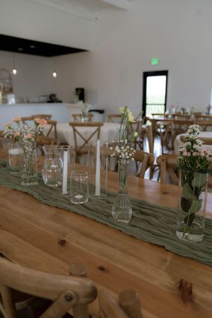Wedding reception table setup with glass vases, green runners and wooden cross-back chairs.