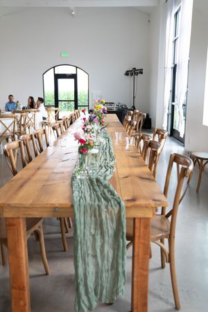 A rustic wedding reception table with wooden chairs and sage green runner decorated with floral arrangements.