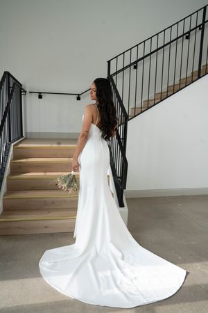 Bride in flowing white wedding dress poses elegantly near wooden staircase.