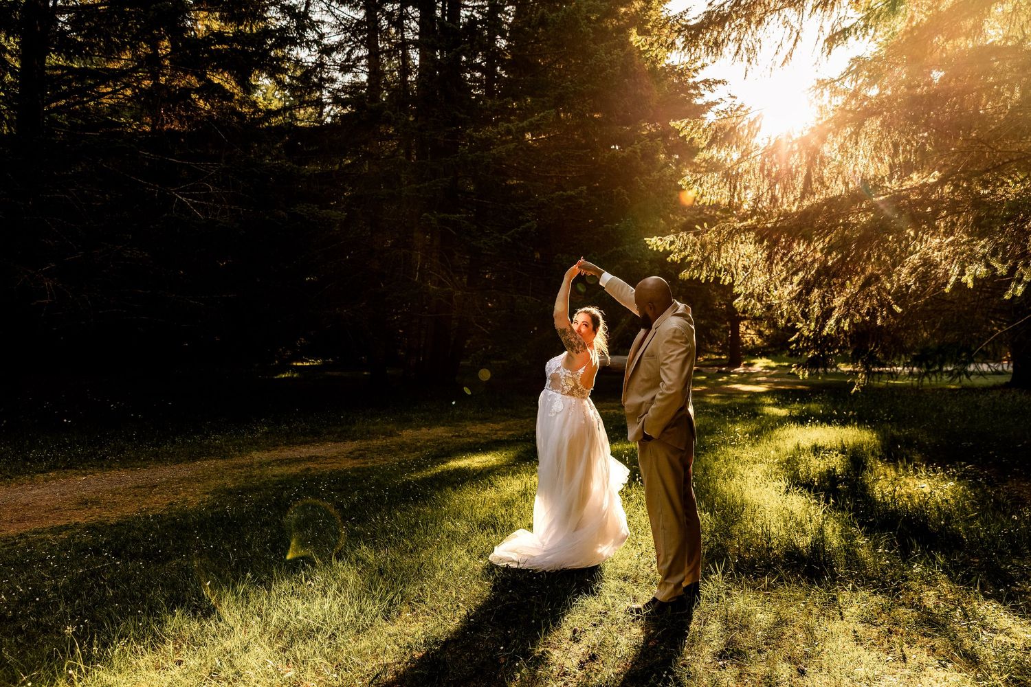 A couple dances together in a sun-drenched forest clearing during golden hour, with light streaming through the trees.
