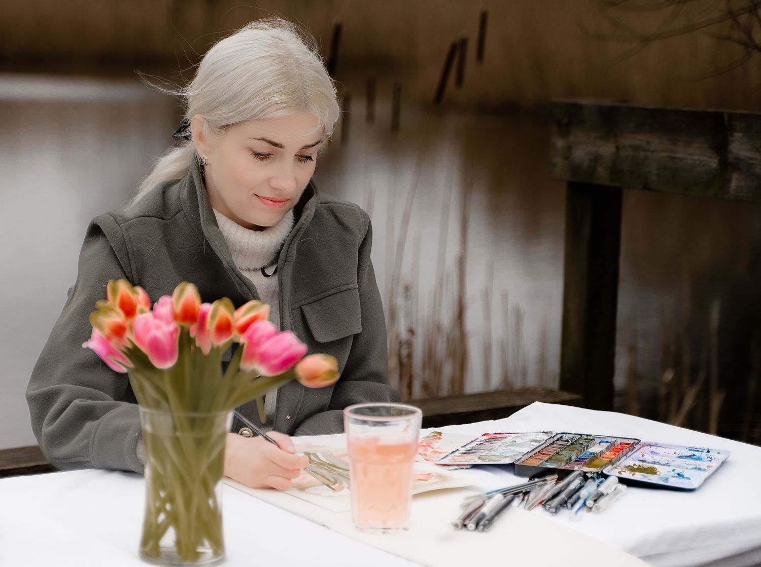 A female artist sits peacefully at an outdoor table near a pond, painting with watercolours beside a vase of tulips and a glass of pink lemo