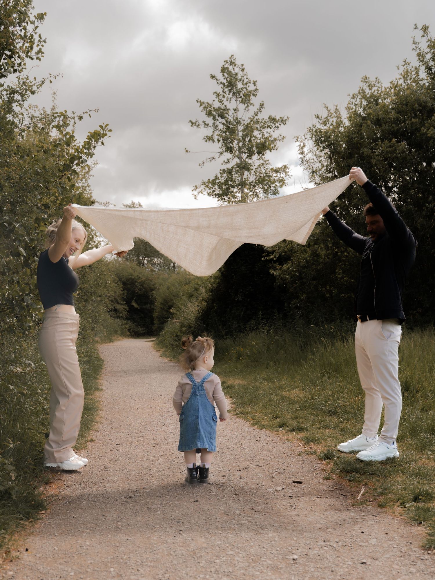 Parents lifting a light blanket above their daughter on a forest path, creating a moment of play and connection during a relaxed family sess