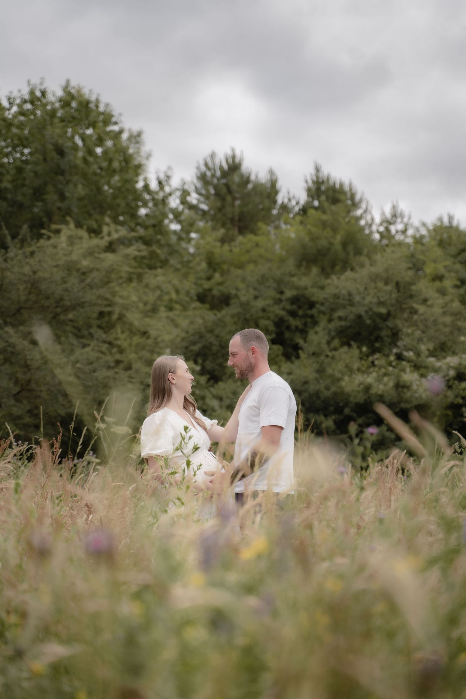 A couple standing close together in a wildflower-filled field, sharing a quiet moment during a maternity session surrounded by soft summer g