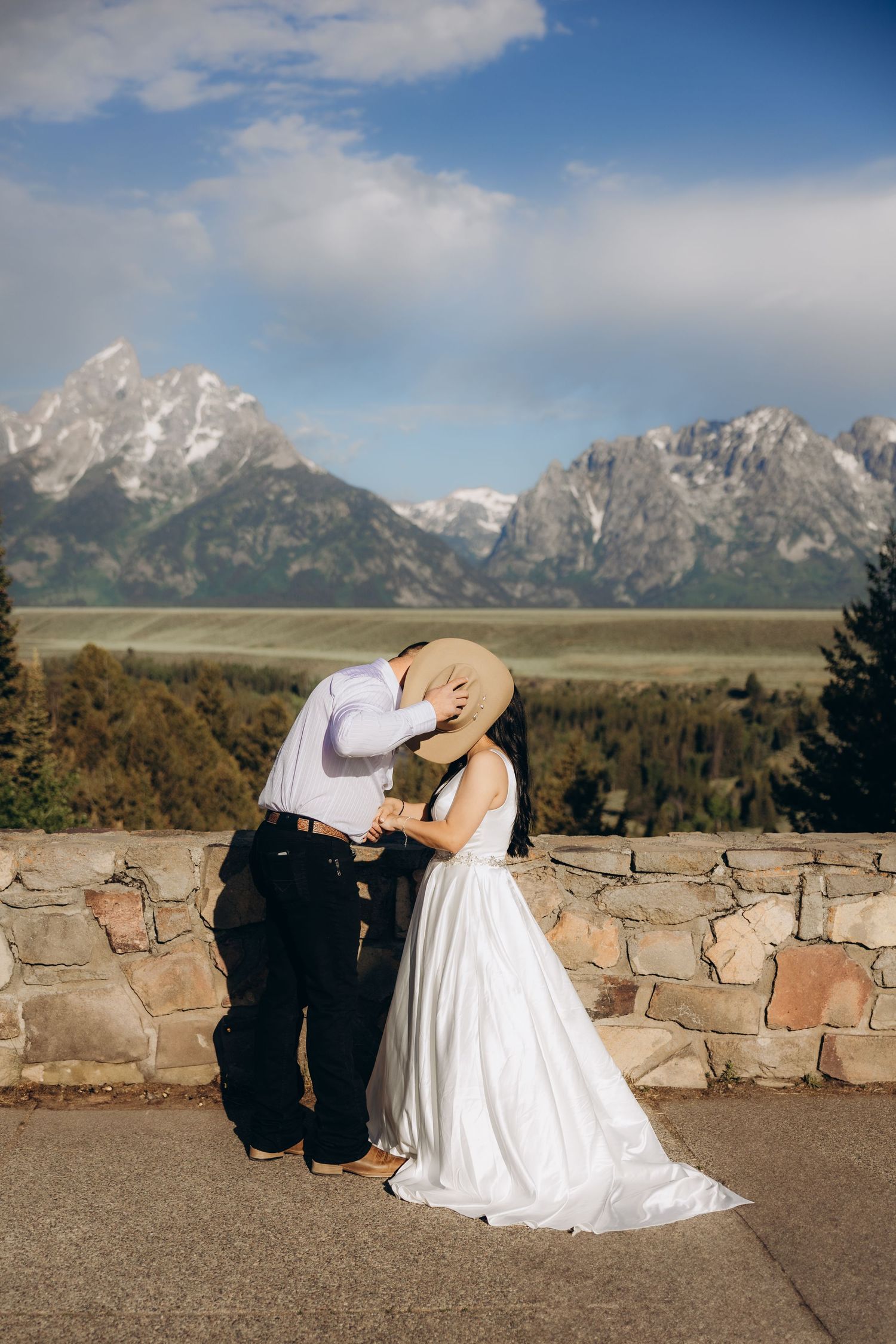 A couple sits together on a stone wall overlooking the majestic Grand Teton mountains at scenic overlook in Wyoming.