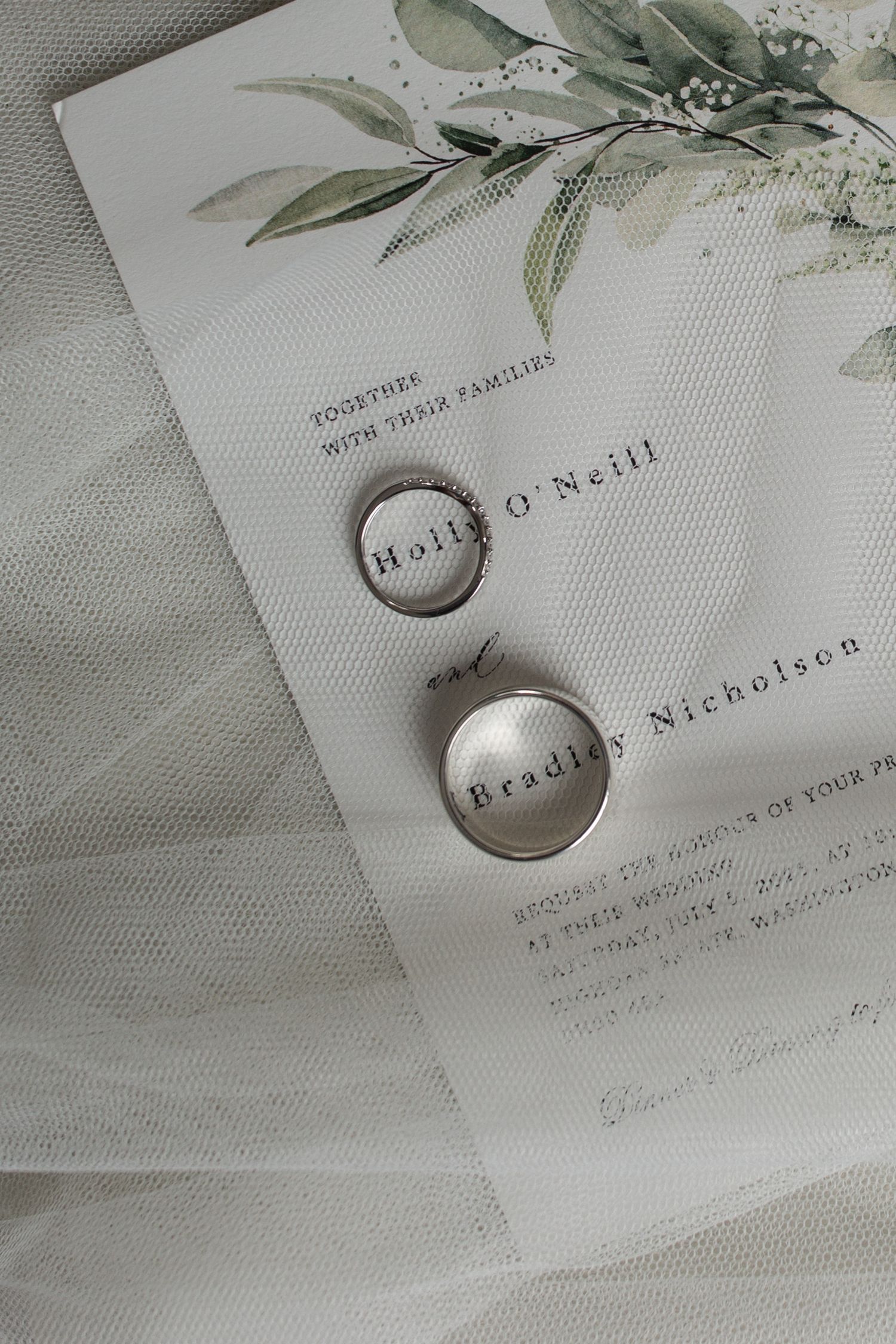 Wedding rings and invitation card with delicate green watercolor botanical details on light textured background.