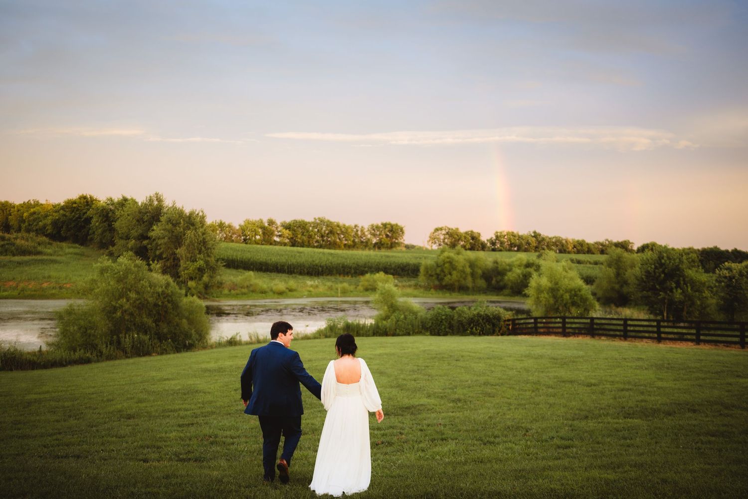 A couple in wedding attire walks through a scenic green meadow at sunset with trees in the distance.