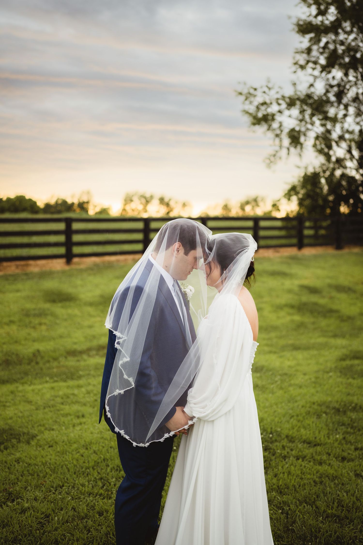 A romantic wedding photo at sunset with the couple standing together on a lush green lawn with wooden fences in background.