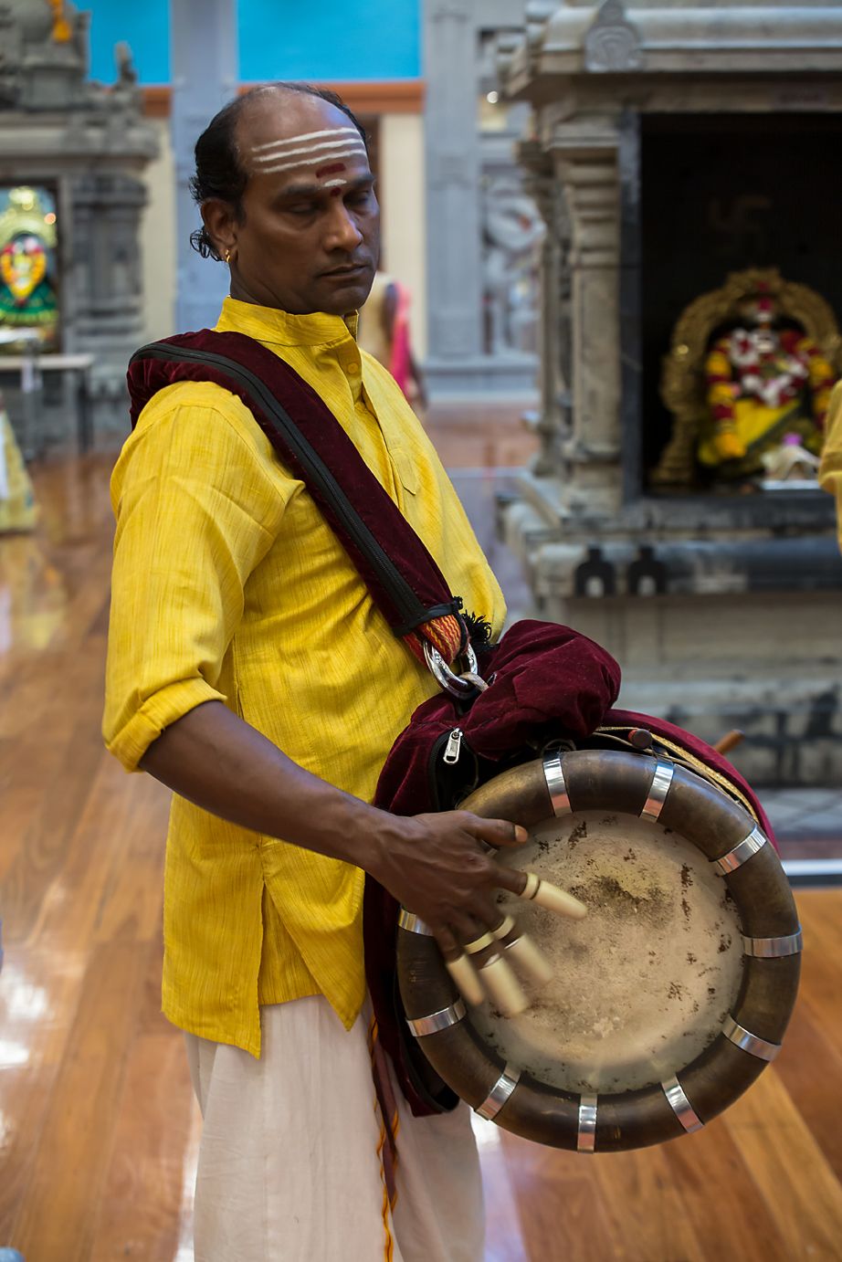 A musician in a yellow shirt plays a traditional temple drum during a performance in a Hindu temple interior.