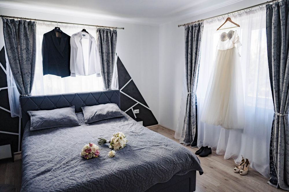 A bedroom interior with gray bedding, patterned curtains, and formal attire hanging on display.
