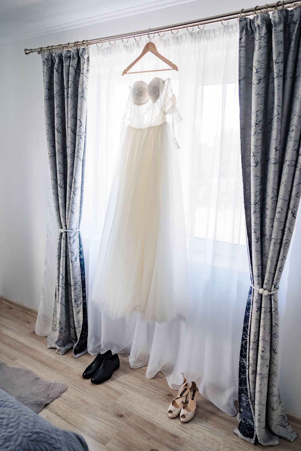 Wedding dress hangs elegantly by window with gray and white curtains.