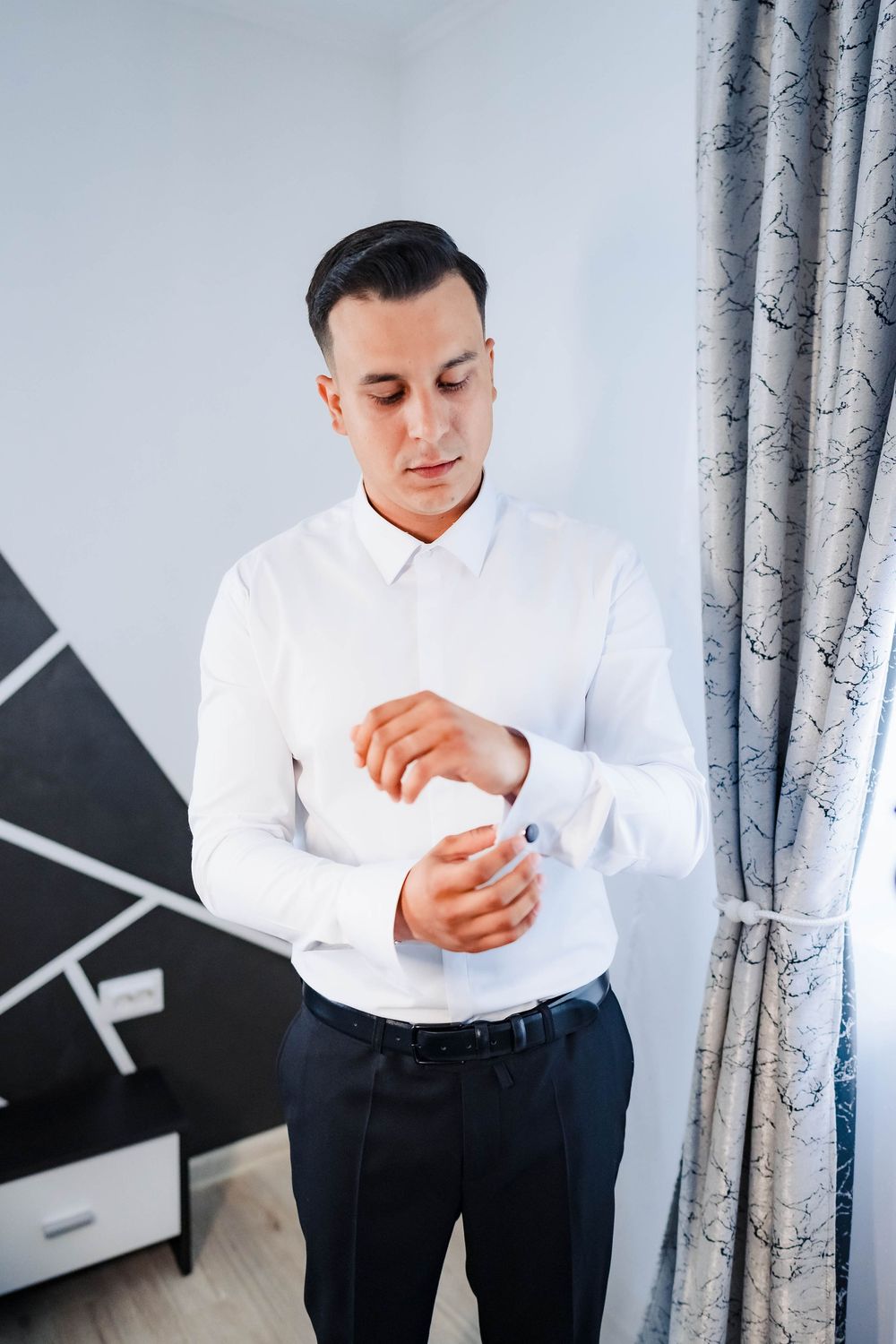 A groom in a white dress shirt adjusts his cuffs while getting ready in a bright room with patterned curtains.