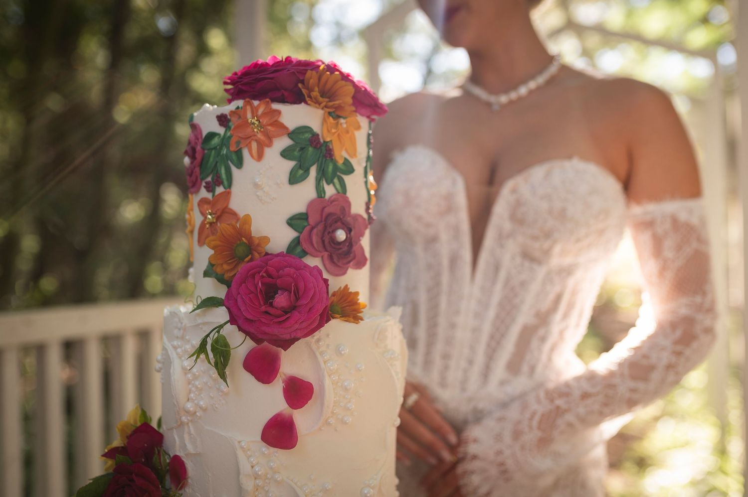 A white tiered wedding cake decorated with vibrant pink and orange flowers sits in front of a lace dress and sunlit backdrop.