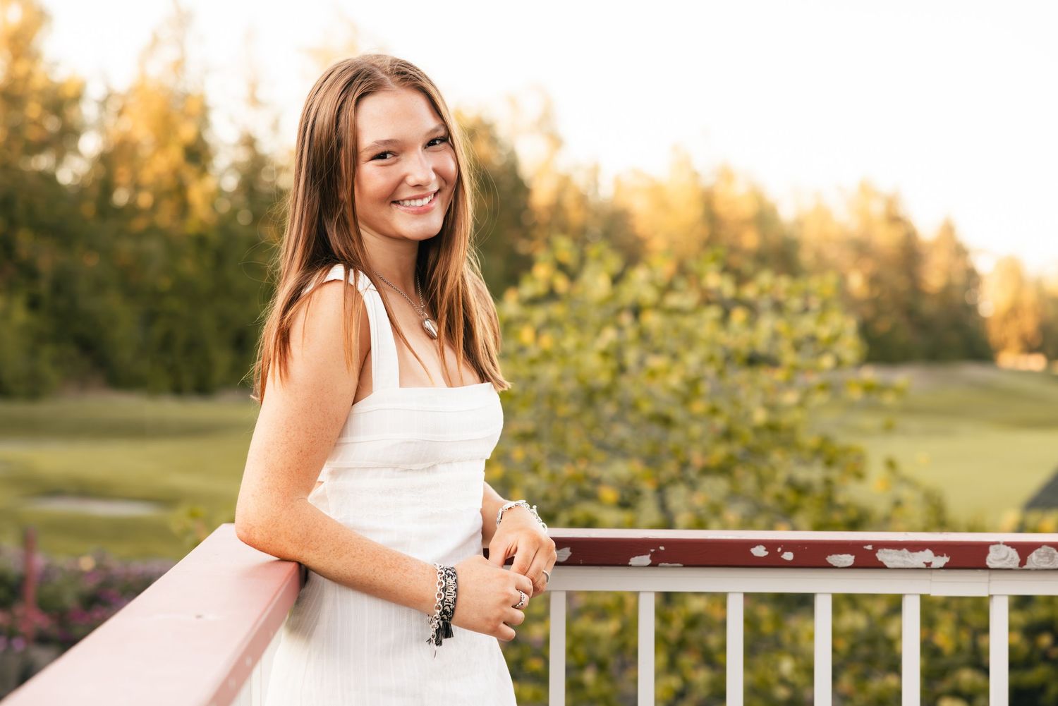 Woman in white dress smiles while leaning on railing as golden sunset illuminates outdoor garden setting.