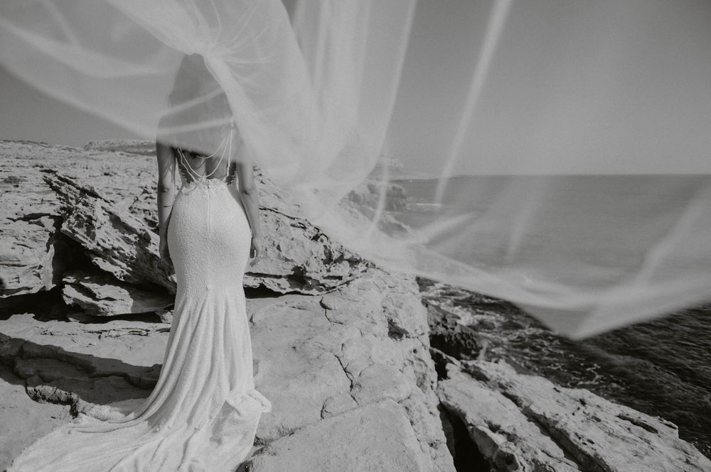 Flowing wedding dress and veil flutter in wind against rocky coastal backdrop.