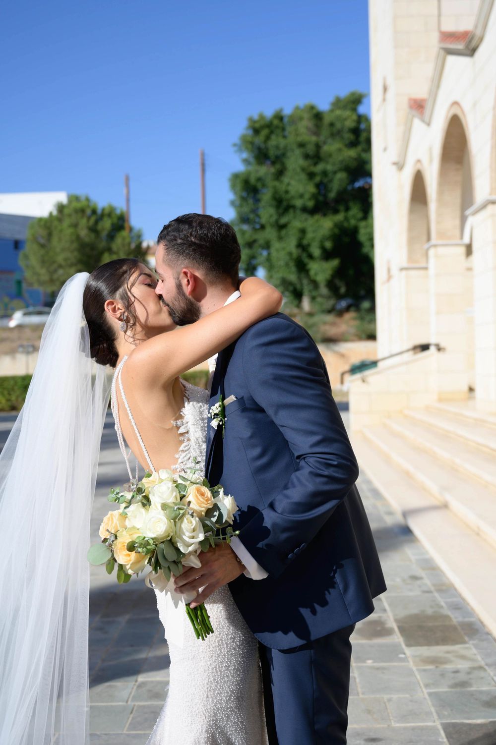 Bride in white lace dress holding bouquet kisses groom in navy suit outside church with arches.