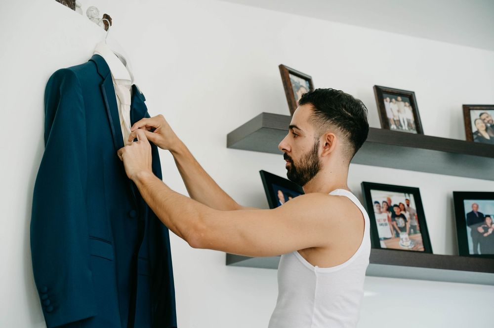 Person in white tank top adjusts formal navy suit on hanger near photo wall.
