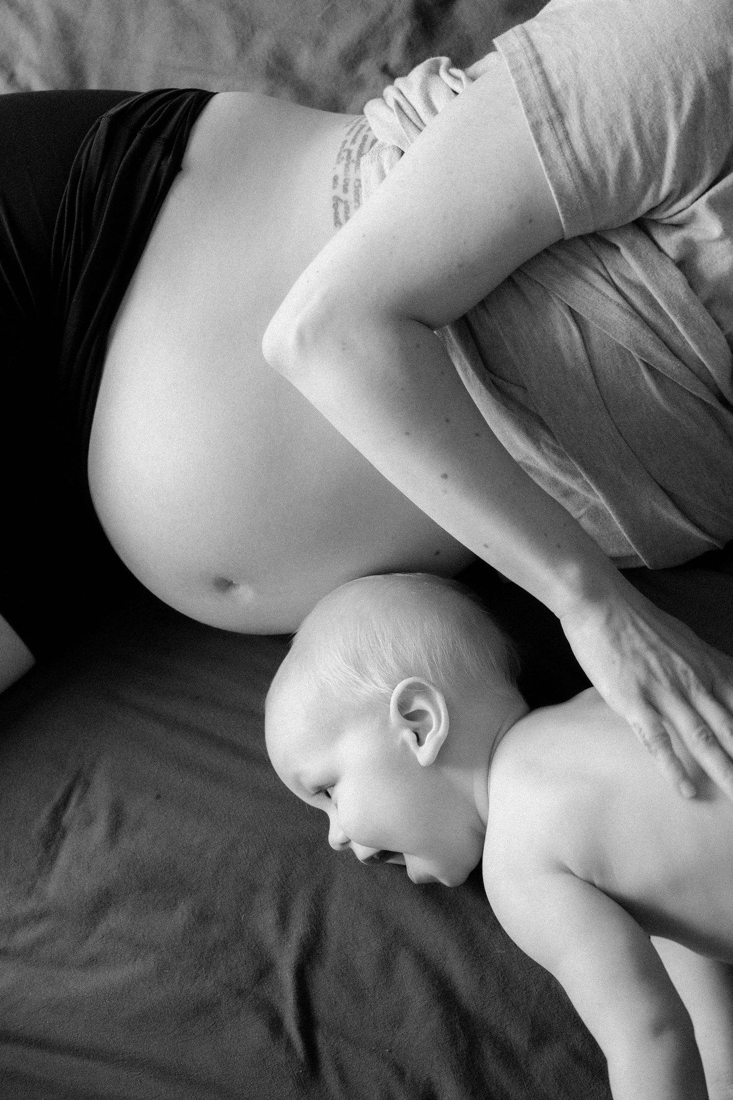 A sleeping newborn baby lies next to a pregnant belly in an intimate black and white photograph.