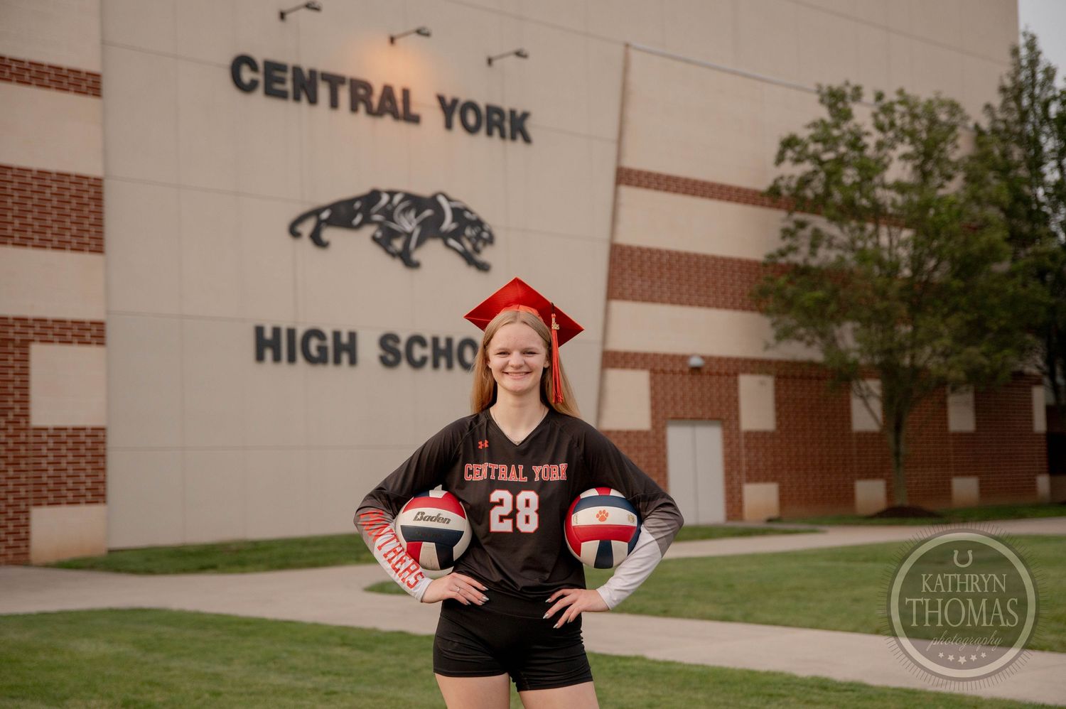 High school baseball player wearing black jersey and red hat poses against brick wall exterior for senior portrait.