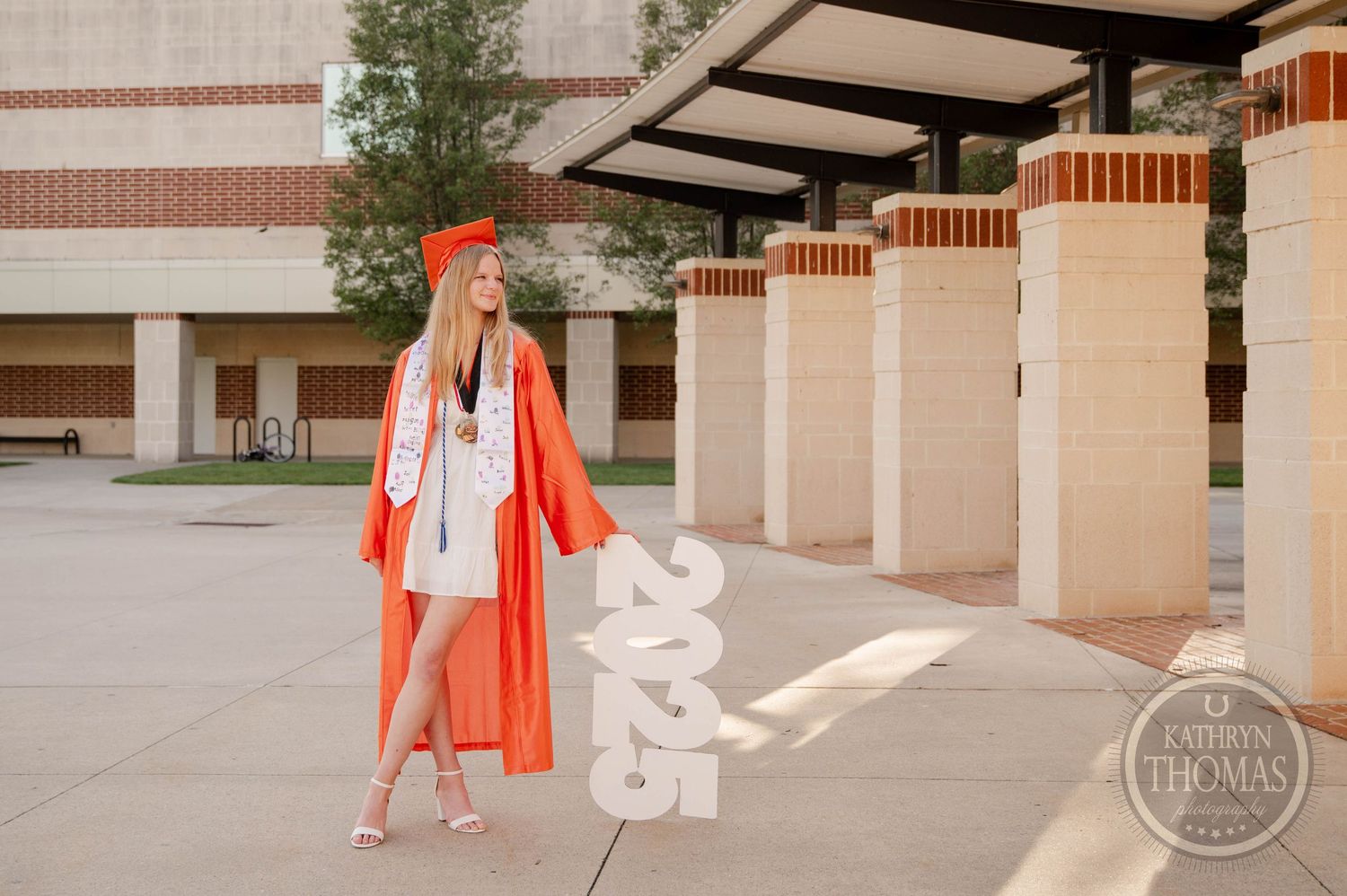 A person in a vibrant orange coat and heels stands in front of modern building columns casting long afternoon shadows.