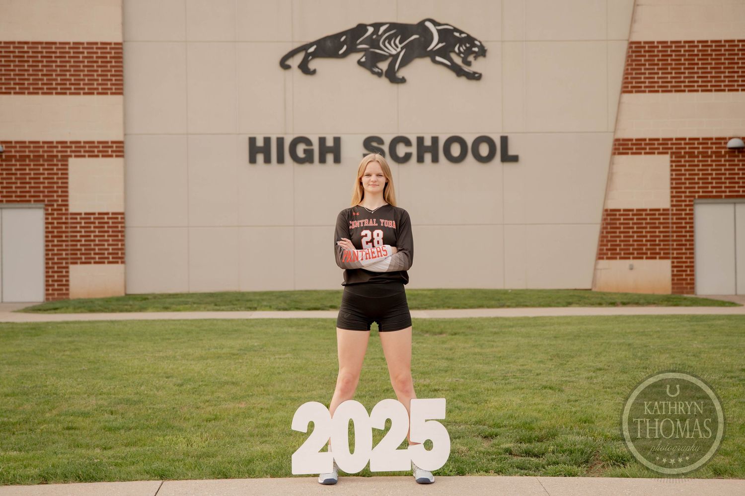 Student wearing black stands on grass in front of high school with panther mascot sign and 2025 numerals.