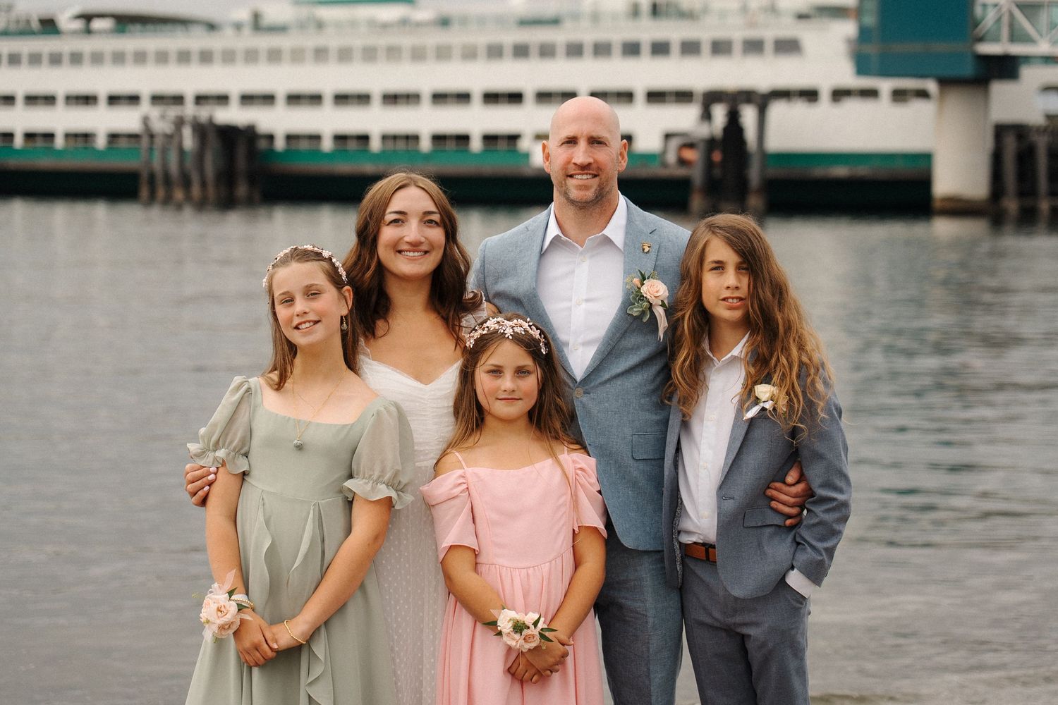 A wedding party in formal attire poses on a dock by the water with boats and buildings visible in the background.