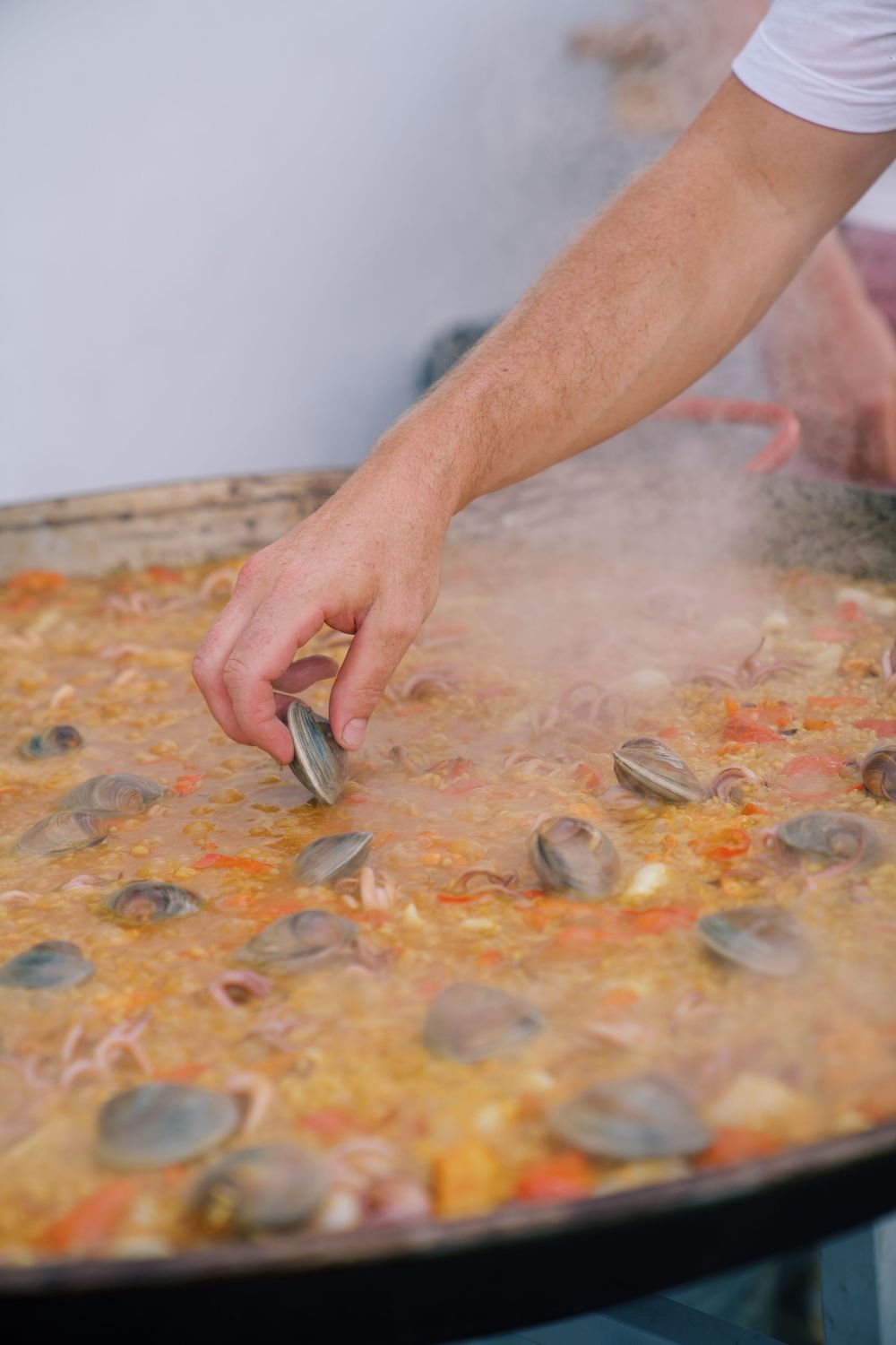 Adding clams to a large paella dish filled with saffron rice and seafood.