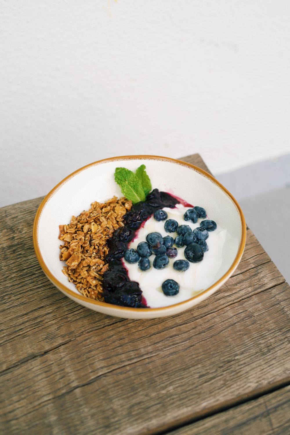 A beautiful breakfast bowl with granola, yogurt, blueberries and mint leaf on wooden table.
