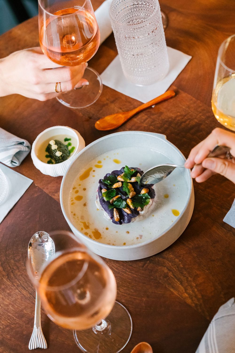Elegant fine dining plate with purple dish topped with greens and nuts, alongside rose wine glasses on wooden table.