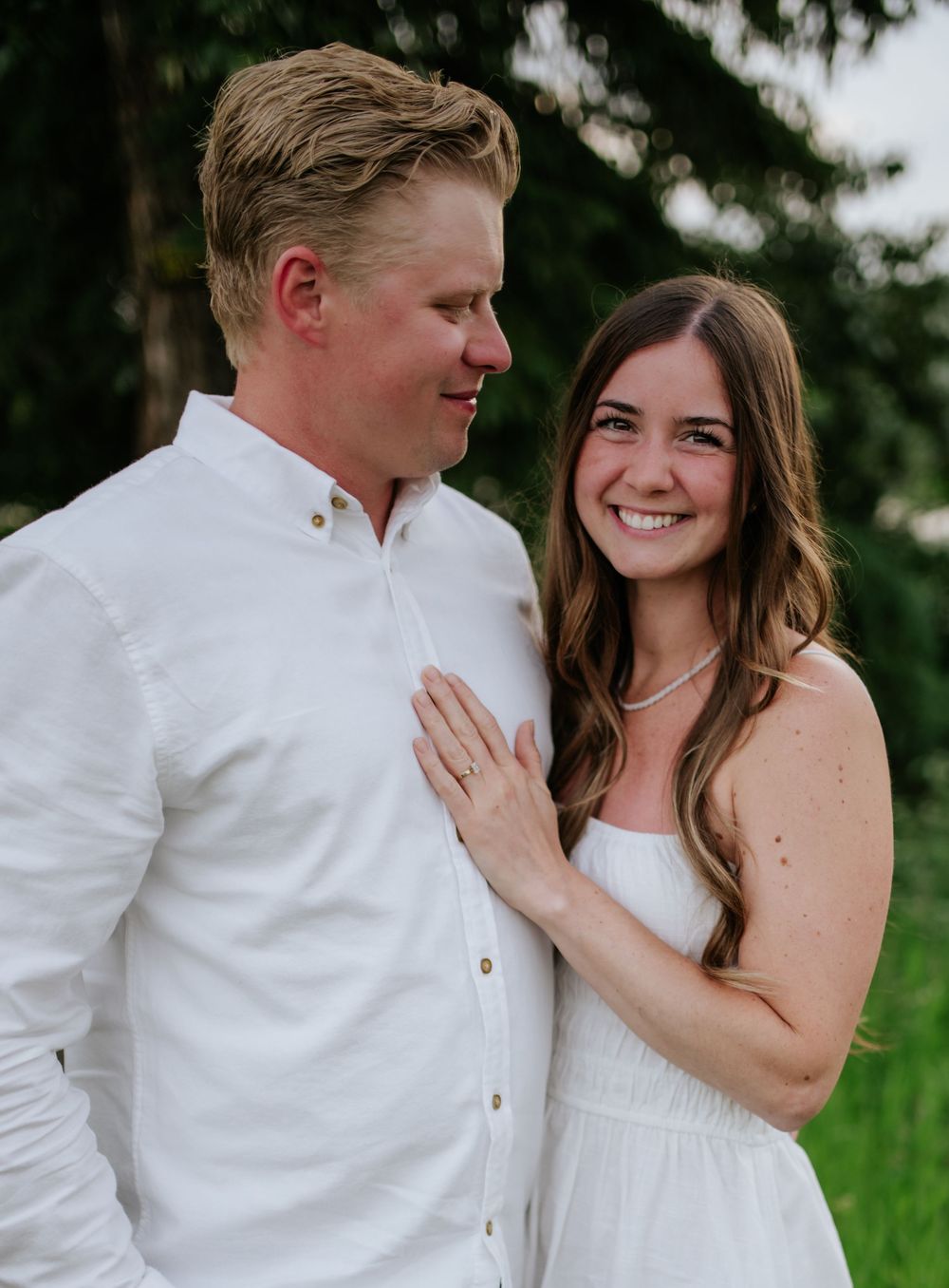 A happy couple poses together outdoors, with the person in white embracing their partner against a natural green background.