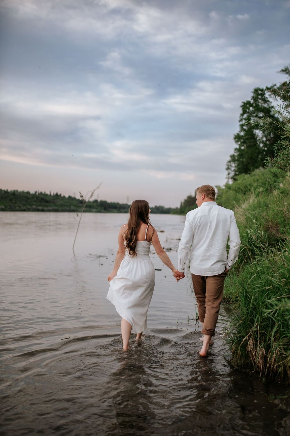 A couple walking hand in hand through shallow river water at sunset with lush greenery along the riverbank.