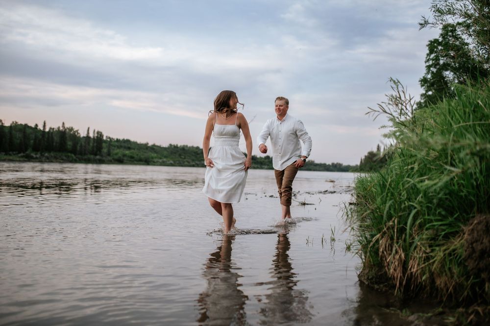 A couple wades joyfully through shallow water at the edge of a serene lake during sunset.