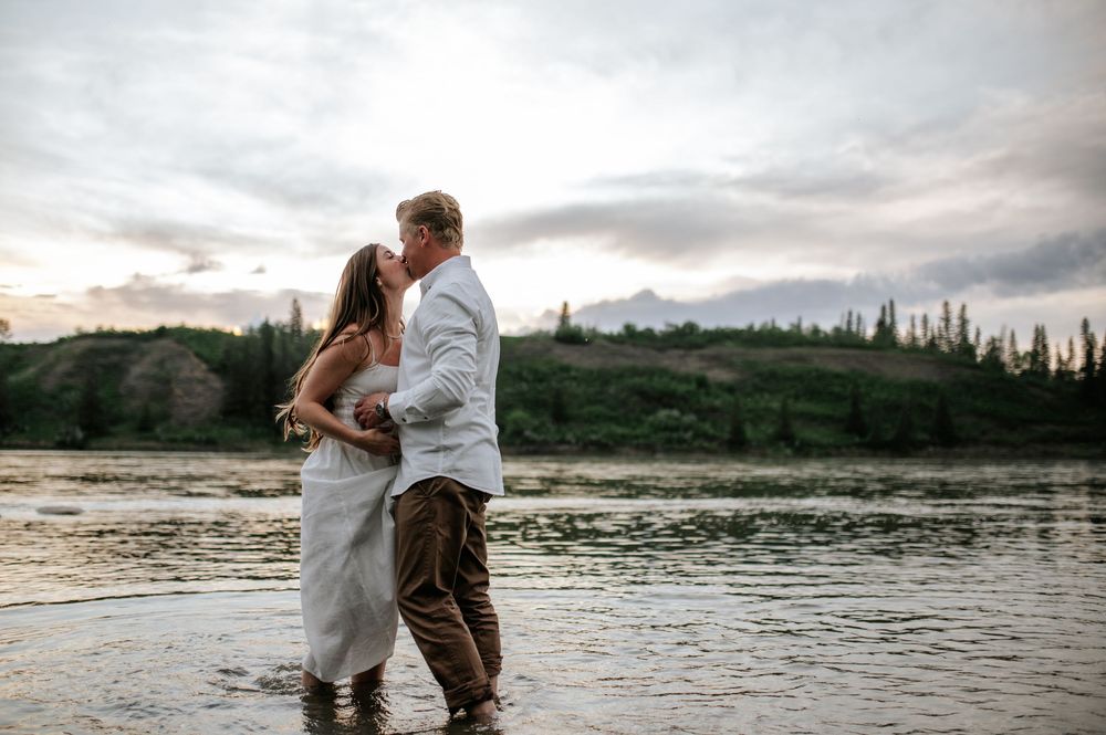 A couple embraces while standing in shallow water at sunset with mountains and trees in the background.
