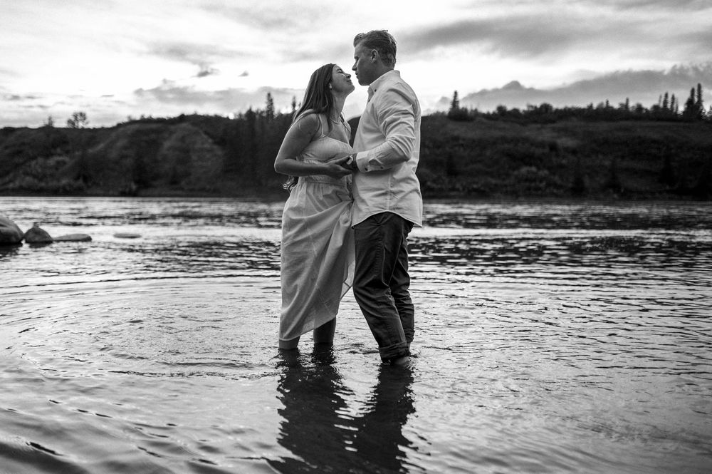 A couple in white clothes stands together in the ocean for beach engagement photos during sunset in black and white.