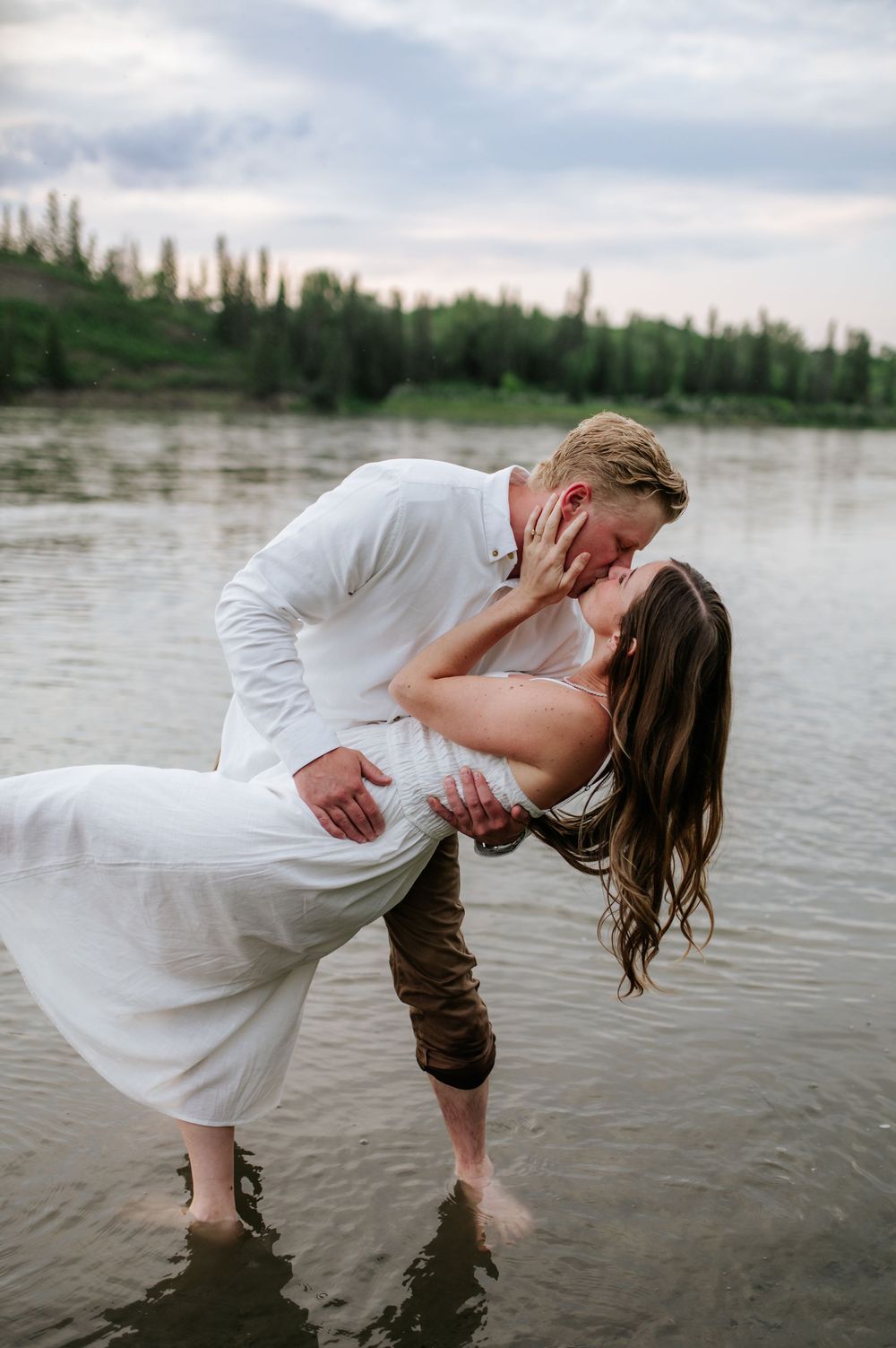 Romantic couple in white clothing embrace and kiss while standing in a scenic lake at sunset.