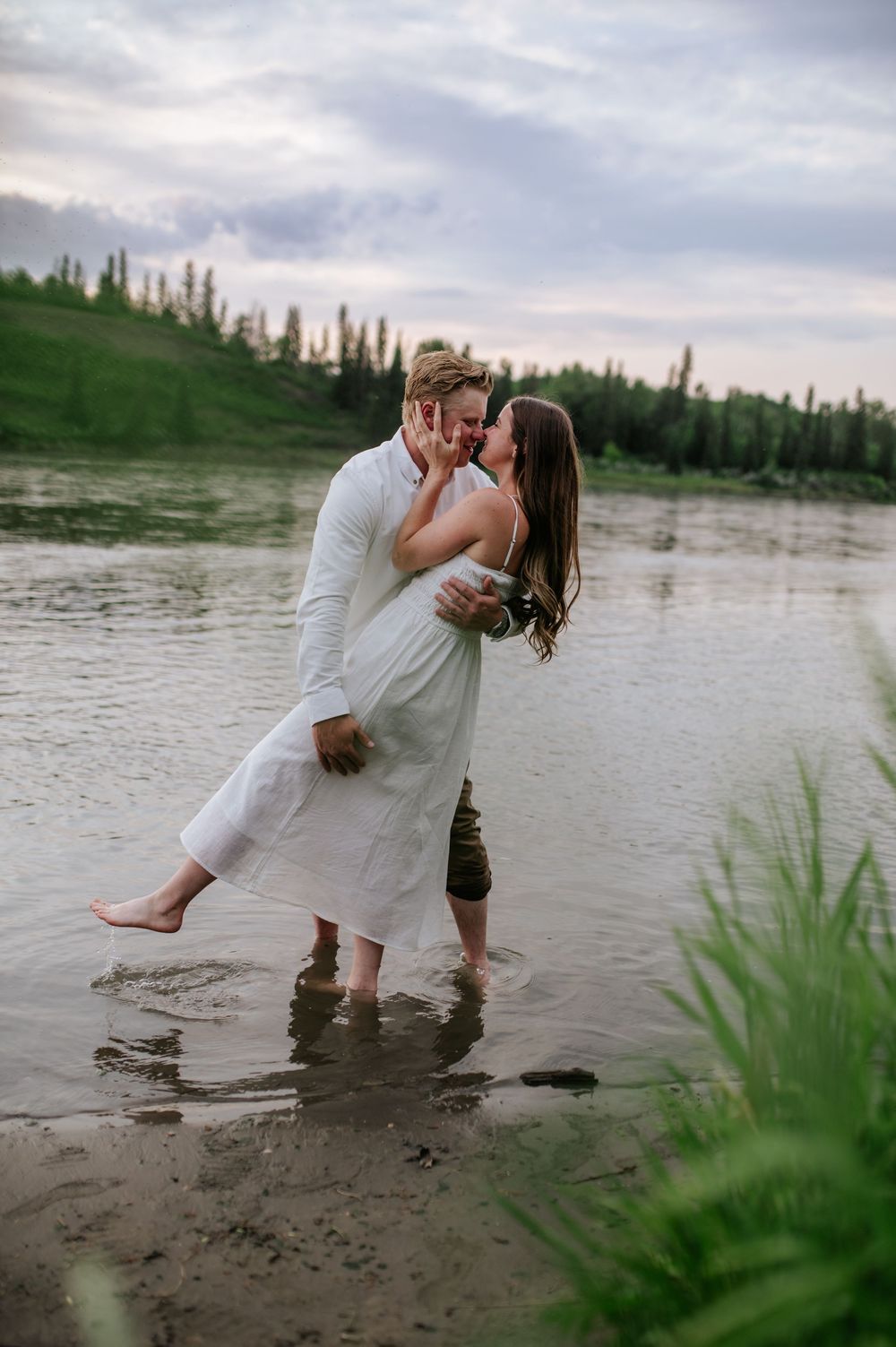 A romantic moment captured by a lake at sunset with two people in white outfits embracing in shallow water.