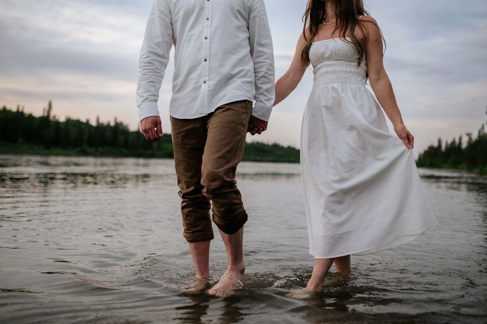 A couple stands together in shallow water at sunset wearing casual white clothing and brown pants.
