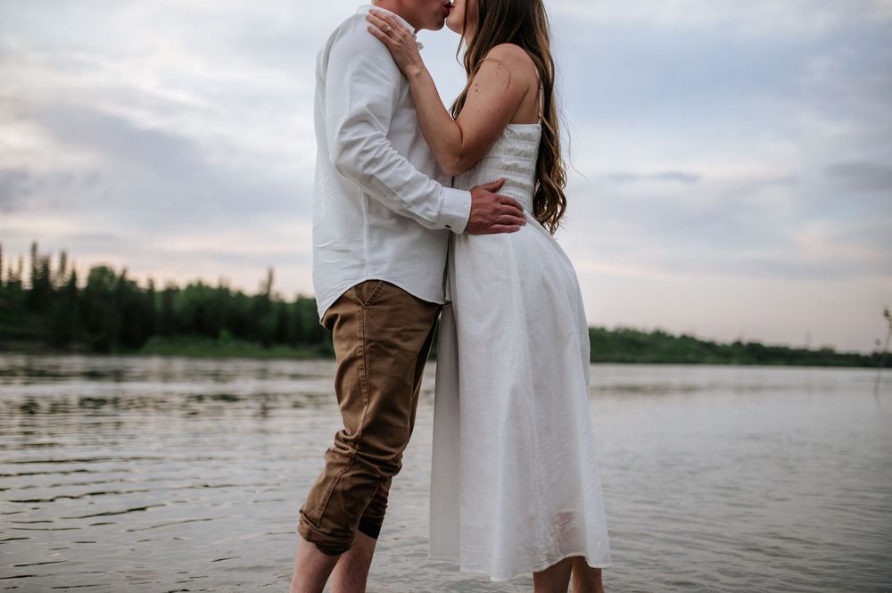 A romantic couple shares an intimate moment by the lake at sunset in white summer attire.