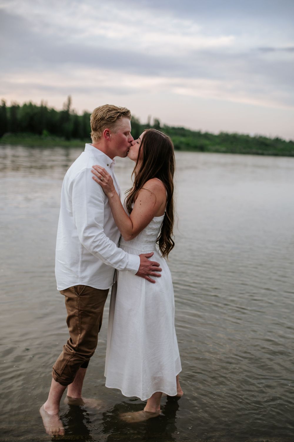 A romantic lakeside couple in white and brown clothing share tender kisses and embraces at sunset.