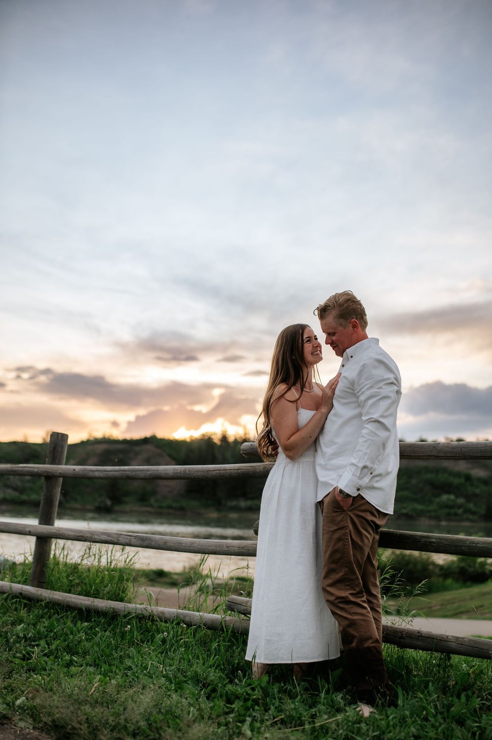 Romantic couple shares intimate moments at sunset by a rustic wooden fence in a scenic countryside setting.