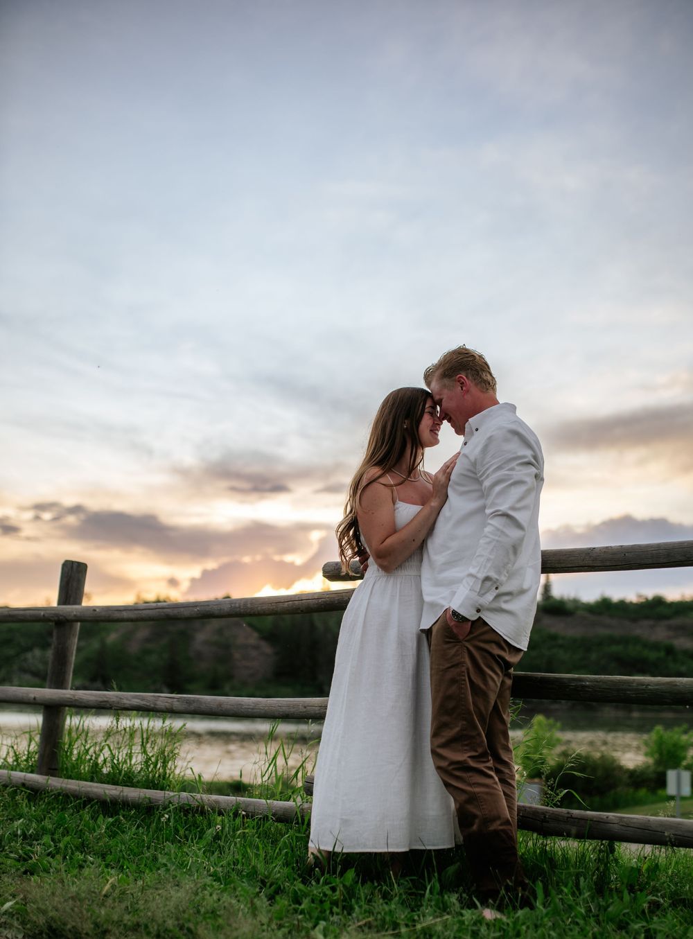 Two people share a romantic moment by a wooden fence at sunset in the countryside.