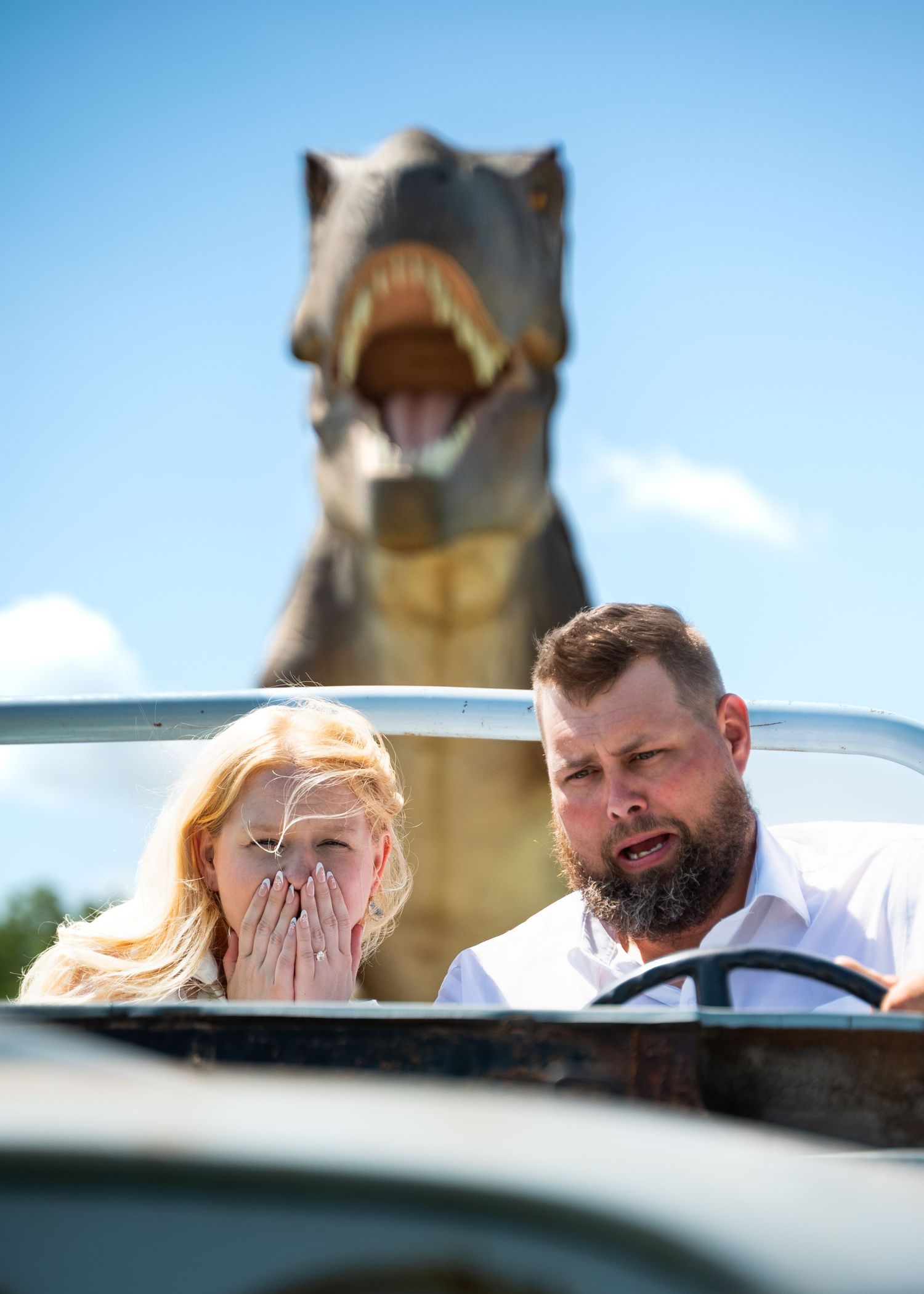 Couple with scared looks on their faces driving a jeep as a T-Rex chases them through Powell Gardens