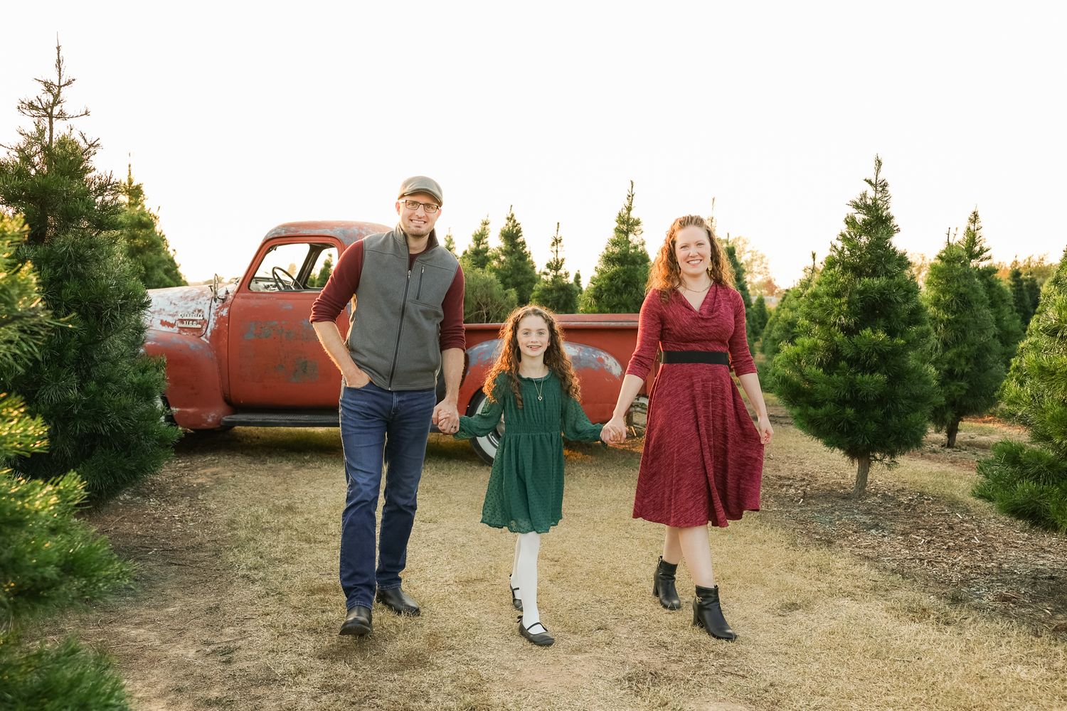A family walks together on a Christmas tree farm with vintage red truck in the background at sunset.