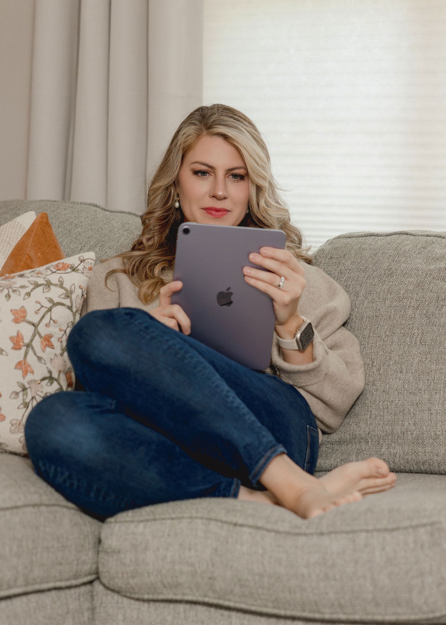 Person relaxing on gray couch using iPad tablet device while wearing casual sweater and jeans.