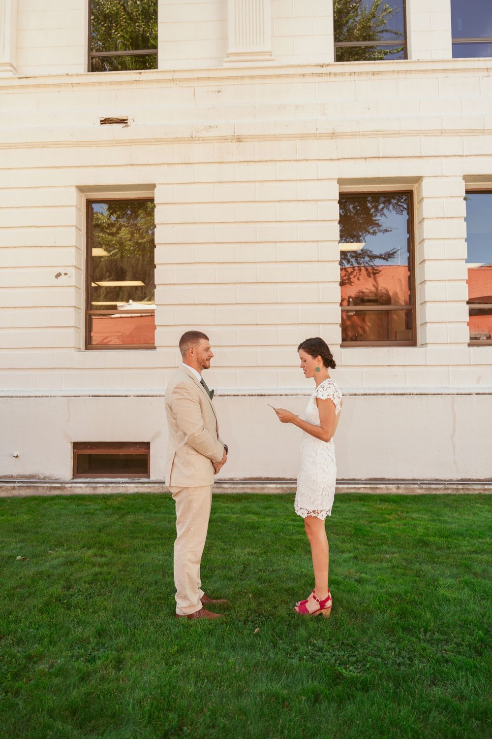 Couple in formal attire shares emotional moment on grass lawn by white building.
