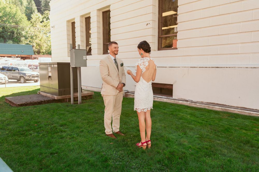 A couple in formal attire stands on a green lawn outside a white building on a sunny day.