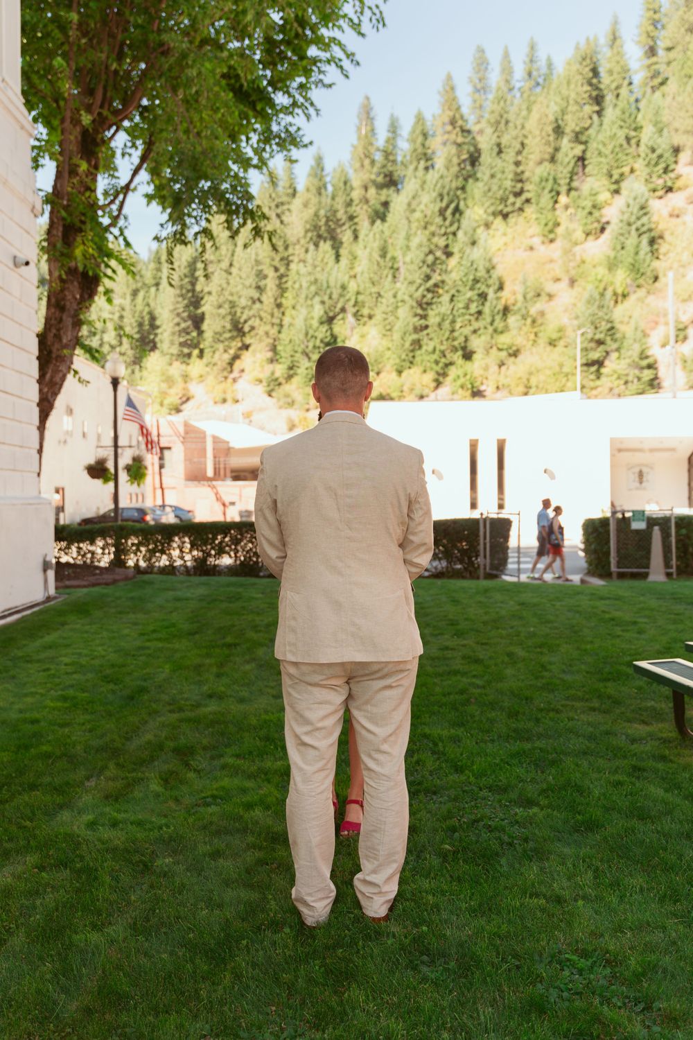 Person in beige suit stands alone on green lawn with pine trees in background.